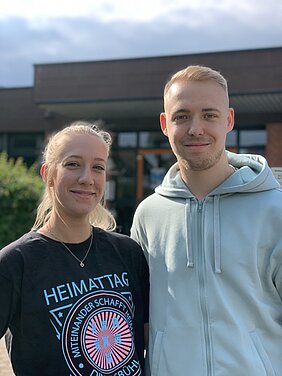 Zwei junge Menschen stehen draußen vor einem Gebäude. Die Frau trägt ein schwarzes T-Shirt mit dem Aufdruck "HEIMATTAG", während der Mann einen hellblauen Hoodie trägt. Beide lächeln freundlich in die Kamera.