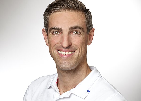 Ein lächelnder Mann mit kurzen, hellen Haaren trägt ein weißes Polohemd. Der Hintergrund ist hell und neutral, was den Fokus auf sein freundliches Gesicht lenkt.