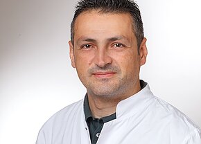 Portrait eines Mannes in einem weißen Kittel. Er hat kurzes, dunkles Haar und einen freundlichen Gesichtsausdruck. Der Hintergrund ist hell und unscharf, wodurch der Fokus auf seinem Gesicht liegt.