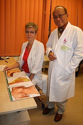 Zwei Ärzte in weißen Kitteln stehen an einem Tisch. Auf dem Tisch befindet sich ein anatomisches Poster, das den menschlichen Körper zeigt. Der Arzt auf der rechten Seite schaut direkt in die Kamera, während die Ärztin links konzentriert auf das Poster blickt.