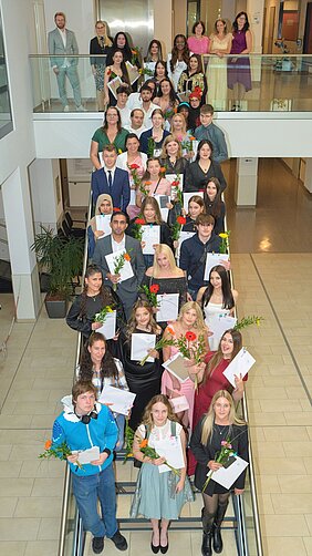 Eine große Gruppe von Absolventen steht auf einer Treppe, einige halten Urkunden und Blumen in den Händen. Der Raum ist hell erleuchtet, und viele Teilnehmer sind feierlich gekleidet. Die Stimmung wirkt festlich und stolz.