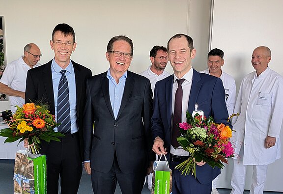 Drei Männer in Anzügen stehen im Vordergrund, jeder mit einem Strauß Blumen in der Hand. Sie lächeln freundlich. Im Hintergrund sind mehrere Personen in weißen Kitteln sichtbar, die ebenfalls lächeln. Die Szene vermittelt eine festliche und herzliche Atmosphäre.