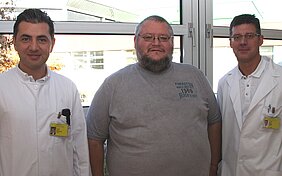 Drei Männer stehen zusammen vor einem Fenster. Zwei tragen weiße Arztkittel, während einer in einem grauen T-Shirt gekleidet ist. Sie wirken freundlich und lächeln, was auf eine positive Atmosphäre hindeutet.