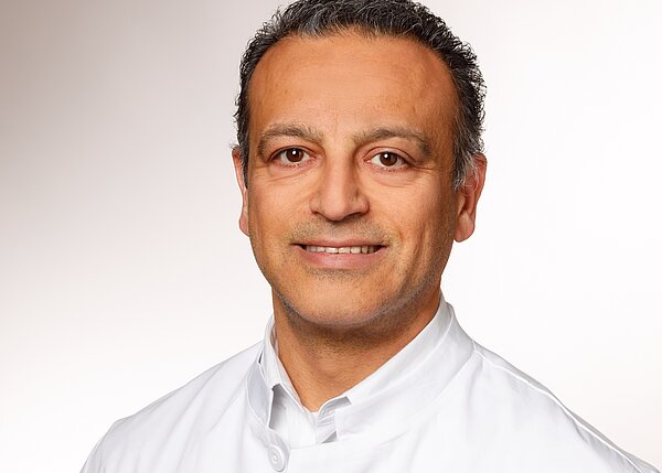 Ein lächelnder Mann trägt einen weißen Kittel. Sein Gesicht ist freundlich und er hat dunkle, leicht wellige Haare. Der Hintergrund ist schlicht und hell. Die Darstellung vermittelt einen professionellen Eindruck, der häufig mit medizinischen Berufen assoziiert wird.