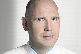 Nahaufnahme eines Mannes mit Glatze und kurzem Haar, der direkt in die Kamera schaut. Er trägt ein helles Hemd und hat einen freundlichen, neutralen Ausdruck auf seinem Gesicht. Im Hintergrund sind verschwommene, helle Linien zu sehen.