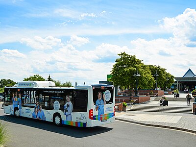 Ein Bus mit Werbung für eine Karriere im Gesundheitswesen fährt vorbei. Auf der Seite des Busses sind mehrere Personen in medizinischer Kleidung abgebildet. Der Himmel ist blau mit weißen Wolken, und im Hintergrund sind Gebäude und Bäume sichtbar. anfahrt_stadtbus_LUX_7090.jpg