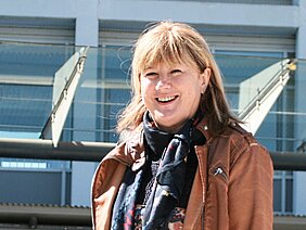 Eine lächelnde Frau steht im Freien, trägt eine braune Jacke und einen Schal. Im Hintergrund sind moderne Gebäude mit großen Fenstern sichtbar. Die Szene vermittelt eine freundliche und entspannte Atmosphäre.