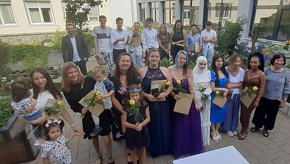 Eine Gruppe von Menschen steht in einem Garten, viele halten Blumen und Urkunden in den Händen. Einige Kinder sind ebenfalls dabei. Es scheint eine Feier zu sein, möglicherweise zur Ehrung oder Graduierung. Im Hintergrund sind Pflanzen und Gebäude sichtbar.