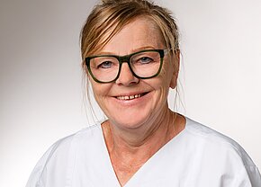 Eine lächelnde Frau mit blonden Haaren trägt eine weiße Arbeitskleidung und grüne Brille. Ihr Gesichtsausdruck vermittelt Freundlichkeit und Professionalität, was auf einen Gesundheitsberuf hinweisen könnte. Der Hintergrund ist neutral und unauffällig, um den Fokus auf die Person zu lenken.