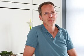 Ein Mann mit brünettem, kurzem Haar und einer Brille sitzt mit einem nachdenklichen Ausdruck. Er trägt ein beige-blaues Polohemd. Im Hintergrund sind Pflanzen sichtbar. Die Umgebung wirkt hell und freundlich.