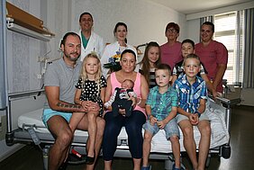 Eine Mutter sitzt im Krankenhausbett und hält ein neugeborenes Baby. Um sie herum stehen Familienmitglieder und Krankenhauspersonal. Die Atmosphäre ist festlich, mit einigen Blumen im Hintergrund. Die Personen wirken glücklich und zufrieden, was auf einen besonderen Anlass hindeutet.