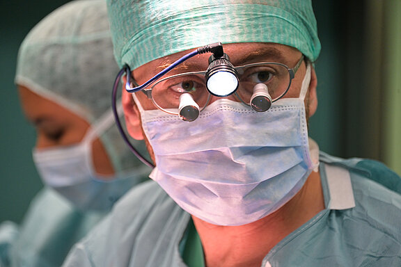Ein Chirurg mit einer grünen OP-Kappe und einer Schutzmaske sieht konzentriert aus. Er trägt eine Brille mit integrierter Beleuchtung. Im Hintergrund ist eine andere Person in OP-Kleidung zu erkennen. Die Szene vermittelt den Eindruck einer chirurgischen Umgebung.