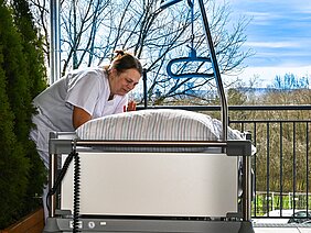 Eine Pflegerin beugt sich über ein Bett mit einem gestreiften Kissen. Im Hintergrund sind Bäume und ein klarer Himmel zu sehen. Die Szene vermittelt eine ruhige, fürsorgliche Atmosphäre.