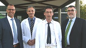 Vier Männer stehen vor einem modernen Gebäude. Drei von ihnen tragen weiße Laborkittel, während einer ein Anzugshirt und eine Krawatte trägt. Die Gruppe wirkt freundlich und professionell. Im Hintergrund sind unscharfe Details der Umgebung sichtbar.