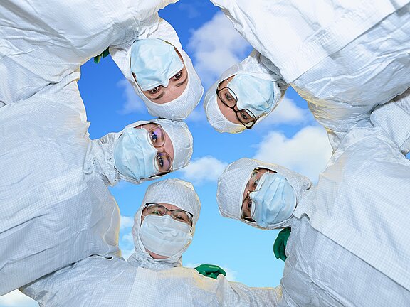 Fünf Personen in weißer Schutzkleidung und Masken blicken lächelnd in die Kamera. Sie stehen in einem Kreis, vor einem blauen Himmel mit Wolken. Der Ausdruck vermittelt Teamgeist und Optimismus in einem medizinischen oder wissenschaftlichen Kontext.