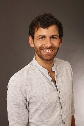 Ein lächelnder Mann mit kurzer, lockiger, dunkler Haarpracht und einem Bart steht vor einem neutralen Hintergrund. Er trägt ein helles, lässiges Hemd mit Knopfleiste. Der Ausdruck seines Gesichts vermittelt Freundlichkeit und Offenheit.