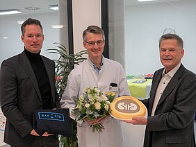 Drei Männer stehen in einem Raum. Einer trägt einen weißen Kittel und hält einen Blumenstrauß, während ein anderer eine Torte mit dem Logo einer Organisation präsentiert. Der dritte Mann hält ein Geschenk. Alle lächeln und wirken erfreut.