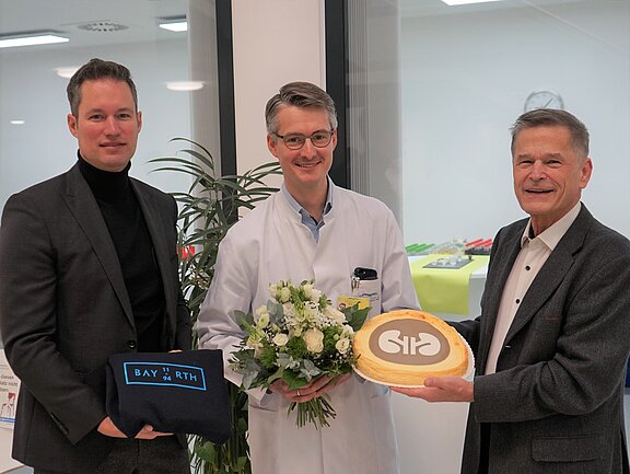 Drei Männer stehen in einem Raum. Einer trägt einen weißen Kittel und hält einen Blumenstrauß, während ein anderer eine Torte mit dem Logo einer Organisation präsentiert. Der dritte Mann hält ein Geschenk. Alle lächeln und wirken erfreut.