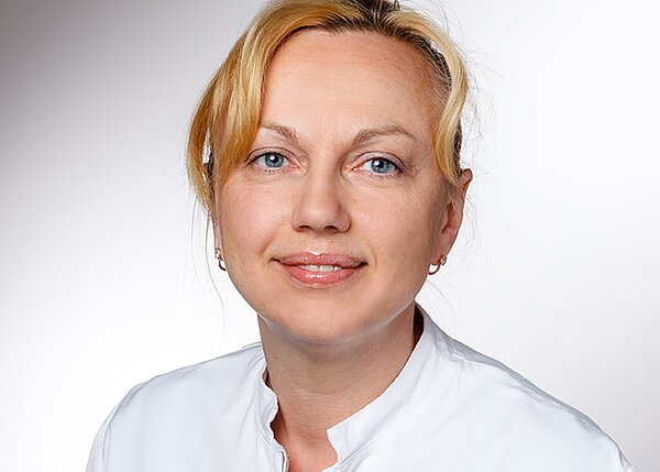 Ein Lächeln einer Frau mit blonden Haaren, die in einem weißen Laborkittel gekleidet ist. Sie hat blaue Augen und trägt kleine Ohrringe. Der Hintergrund ist hell und neutral gehalten.