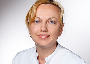 Ein Lächeln einer Frau mit blonden Haaren, die in einem weißen Laborkittel gekleidet ist. Sie hat blaue Augen und trägt kleine Ohrringe. Der Hintergrund ist hell und neutral gehalten.