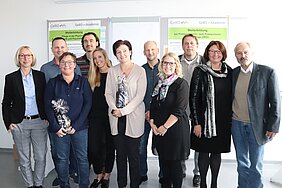 Eine Gruppe von zehn Personen steht zusammen in einem Raum. Sie lächeln und halten Geschenke in den Händen. Im Hintergrund sind Plakate zur Weiterbildung in der Pflege zu sehen. Die Atmosphäre wirkt freundlich und feierlich.