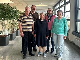 Fünf Personen stehen in einem hellen Raum mit großen Fenstern. Sie lächeln und tragen legere Kleidung. Im Hintergrund sind Pflanzen in Töpfen sichtbar. Die Personen scheinen sich wohlzufühlen und sind freundlich zueinander.