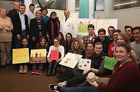 Eine Gruppe von Erwachsenen und Jugendlichen posiert in einem Raum. Sie halten Kunstwerke und Plakate in den Händen, die ihre kreativen Beiträge zeigen. Im Hintergrund sind Zeichnungen von Blumen und Landschaften zu sehen, und zwei Personen in Arztkleidung stehen vorne.