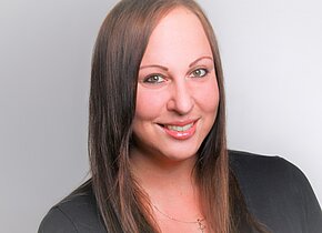 Ein Porträt einer lächelnden Frau mit langen, glatten, braunen Haaren. Sie trägt ein schwarzes Oberteil und eine Halskette mit einem Kreuz. Der Hintergrund ist hell und unscharf, wodurch ihr Gesicht in den Vordergrund tritt.