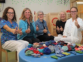 Fünf Frauen sitzen um einen Tisch, auf dem viele gehäkelte Mützen liegen. Alle zeigen mit beiden Daumen nach oben und lächeln. Die Szene vermittelt Freude und Gemeinschaft.