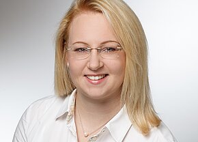 Porträt einer lächelnden Person mit blonden Haaren und einer weißen Bluse. Sie trägt eine dezente Kette mit einer Perle und eine Brille. Der Hintergrund ist hell und neutral, wodurch der Fokus auf dem Gesicht liegt.