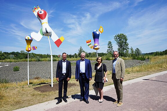 Zwei farbenfrohe Skulpturen stehen im Hintergrund einer Landschaft. Im Vordergrund stehen vier Personen, die sich lächelnd für das Foto positioniert haben. Es ist ein sonniger Tag, und die Umgebung ist grün mit Bäumen und Sträuchern.