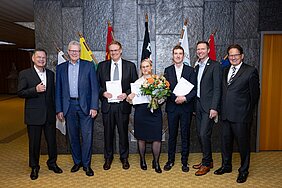 Eine Gruppe von sieben Personen steht in einem Raum, von denen einige Urkunden und eine Blumenstrauß in der Hand halten. Im Hintergrund sind Flaggen zu sehen, was auf eine offizielle Veranstaltung hinweist. Alle wirken erfreut und posieren für ein Foto.