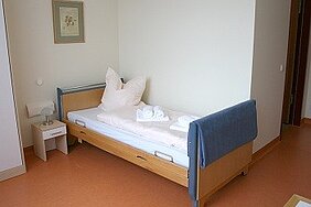 Ein schlichtes, helles Zimmer mit einem Einzelbett. Auf dem Bett liegen Kissen und handtuchartige Gegenstände ordentlich gestapelt. An der Wand hängt ein einfaches Bild. Daneben steht ein Nachttisch mit einer Lampe. Der Boden ist in orangefarbenen Tönen gehalten.