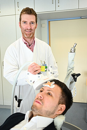 Ein Arzt in einem weißen Kittel hält ein medizinisches Gerät über dem Kopf eines Patienten, der in einem Behandlungsstuhl sitzt. Der Patient wirkt entspannt, während der Arzt sich mit ihm beschäftigt. Im Hintergrund sind medizinische Geräte sichtbar.