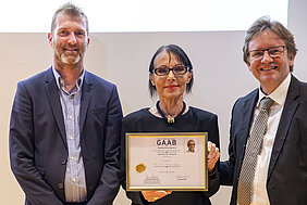 Drei Personen stehen zusammen und halten gemeinsam ein Zertifikat. Die mittlere Person trägt ein schwarzes Outfit und eine elegante Halskette. Das Zertifikat ist ein Erinnerungsdokument, das den Namen „Prof. Dr. Stefan Linder“ trägt und mit dem „GAAB Gedächtnispreis“ ausgezeichnet ist.