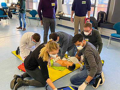 Eine Gruppe von Menschen praktiziert Erste Hilfe in einem Raum. Mehrere Personen sind um eine Übungspuppe versammelt, während jemand aus einer Tasche medizinisches Equipment holt. Alle tragen Schutzmasken und Handschuhe, um hygienisch zu arbeiten. als-Kurs.jpg