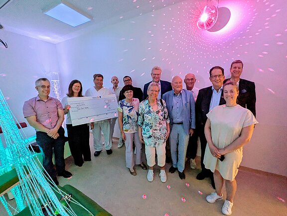 Eine Gruppe von Personen steht in einem hellen Raum, in dem ein großer Scheck präsentiert wird. Die Menschen lächeln und posieren zusammen. Im Hintergrund sind farbige Lichter und eine dekorative Wandgestaltung zu sehen. Der Scheck symbolisiert eine Spende für einen guten Zweck.
