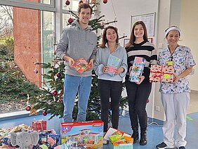 Eine Gruppe von vier Personen steht vor einem Weihnachtsbaum. Sie halten Geschenkpakete in den Händen und jede Person ist lächelnd. Um sie herum liegen viele weitere Geschenke auf dem Boden, die ebenfalls festlich verpackt sind. Im Vordergrund ist ein Plakat mit dem Logo von AKGM zu sehen.