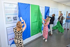 Die Abbildung zeigt eine Gruppe von Personen, die farbige Stoffe (blau und grün) von Informationsständen abziehen. Diese Stände enthalten Forschungsprojekte zur Hybrid-PET-CT-Bildgebung bei nicht kleinzelligem Lungenkrebs. Die Anwesenden wirken interessiert und engagiert.