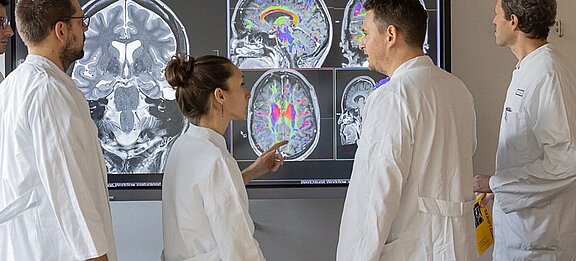 Vier Ärzte in weißen Kitteln stehen vor einem Bildschirm, auf dem Gehirn-MRT-Bilder zu sehen sind. Eine Ärztin zeigt auf bestimmte Bereiche der Bilder, während die anderen aufmerksam beobachten. Die Darstellung ist farbig, um verschiedene Strukturen im Gehirn hervorzuheben.