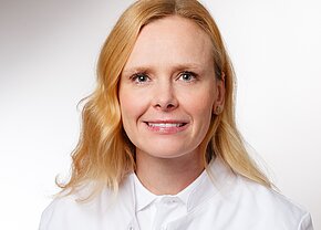 Ein Portrait einer Frau mit blonden, lockigen Haaren, die in einem weißen Arztkittel gekleidet ist. Sie hat ein freundliches Lächeln und schaut direkt in die Kamera. Der Hintergrund ist hell und einheitlich.