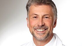 Ein lächelnder Mann mit grauen Haaren und einem Vollbart trägt ein weißes Hemd. Der Hintergrund ist hell und neutral. Der Ausdruck ist freundlich und einladend.
