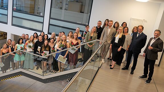 Eine Gruppe von etwa 30 Personen steht auf einer Treppe in einem modernen Gebäude. Sie lächeln und halten Blumen sowie Dokumente in den Händen. Die Anwesenden sind in festlicher Kleidung, was auf einen feierlichen Anlass hinweist.