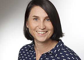 Porträt einer lächelnden Frau mit schulterlangem, dunkelbraunem Haar. Sie trägt ein blaues Hemd mit weißen Schleifenmustern. Der Hintergrund ist hell und unifarben. Ihr Gesichtsausdruck wirkt freundlich und einladend.