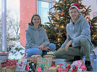 Zwei Personen sitzen neben einem festlich geschmückten Weihnachtsbaum. Um sie herum sind viele mit buntem Geschenkpapier und Schleifen verpackte Geschenke. Die Atmosphäre ist festlich und gemütlich, passend zur Weihnachtszeit. akgm-2022-kinderklinik.jpg