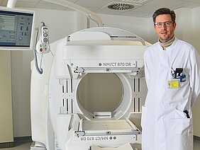Ein Arzt in einem weißen Kittel steht neben einem modernen CT-Gerät. Das Gerät hat einen runden Öffnungsbereich und eine digitale Steuerung. Der Raum ist hell und klinisch. Der Arzt trägt eine Personalausweis-Karte und lächelt freundlich.