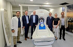 Sechs Personen stehen zur Kamera. Sie befinden sich in einem modernen medizinischen Raum neben einem MRT-Gerät von Siemens. Auf der Liege des Geräts liegen mehrere farbige Luftballons. Der Raum wirkt klinisch und gut beleuchtet, mit neutraler Farbgestaltung.