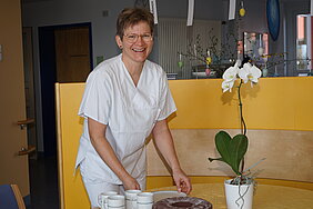 Eine lächelnde Pflegekraft in weißer Arbeitskleidung steht an einem Tisch und serviert Tassen. Neben ihr steht eine blühende Orchidee in einem weißen Topf. Die Umgebung ist hell und freundlich gestaltet.