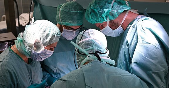 Fünf medizinische Fachkräfte in grünen OP-Kitteln und Mund-Nasen-Schutz konzentrieren sich auf einen chirurgischen Eingriff. Sie stehen um einen Tisch und arbeiten gemeinsam an einem Patienten. Der Raum ist gut beleuchtet, und sterile Instrumente sind sichtbar.