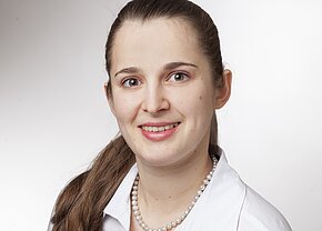 Ein Porträt einer lächelnden Frau mit langen, braunen Haaren, die in einem weißen Hemd und einer Perlenkette gekleidet ist. Sie blickt direkt in die Kamera, mit freundlichen, blauen Augen vor einem hellen Hintergrund.